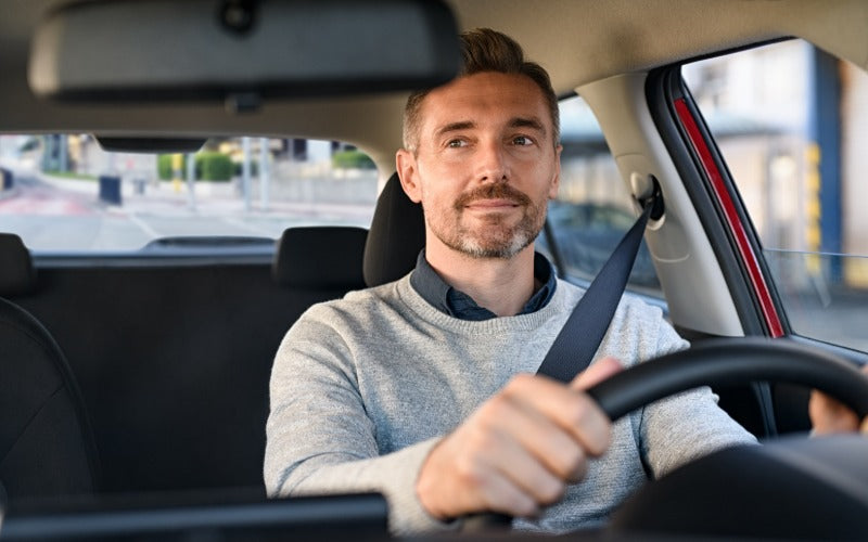 A driver wearing a seatbelt and gray sweater sits behind the wheel, viewed from inside a car on a city street.