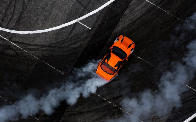 An aerial view of an orange sports car drifting across a dark racetrack, leaving thick tire smoke behind on a sweeping turn.