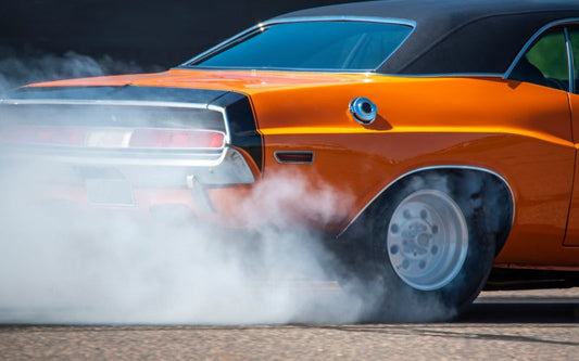 Rear of an orange classic muscle car performing a burnout, with thick white tire smoke billowing around the rear wheel.