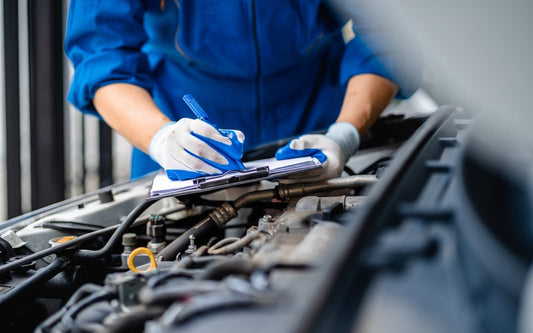 A mechanic in blue coveralls writes on a clipboard above an open engine bay while inspecting vehicle components for service.