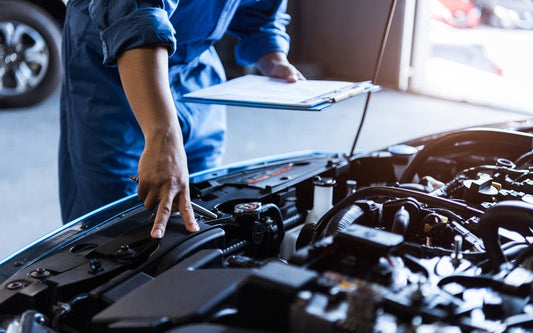 Mechanic in blue coveralls inspects an open car engine, holding a clipboard and pointing into the engine bay.