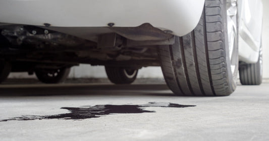 A low angle view of a white car parked in a garage. There is a large oil stain just underneath the back of the car.