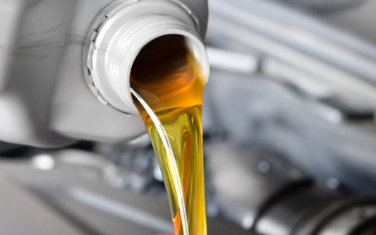 An open wide mouthed gray bottle is pouring thick amber car oil into an open vehicle engine block below.
